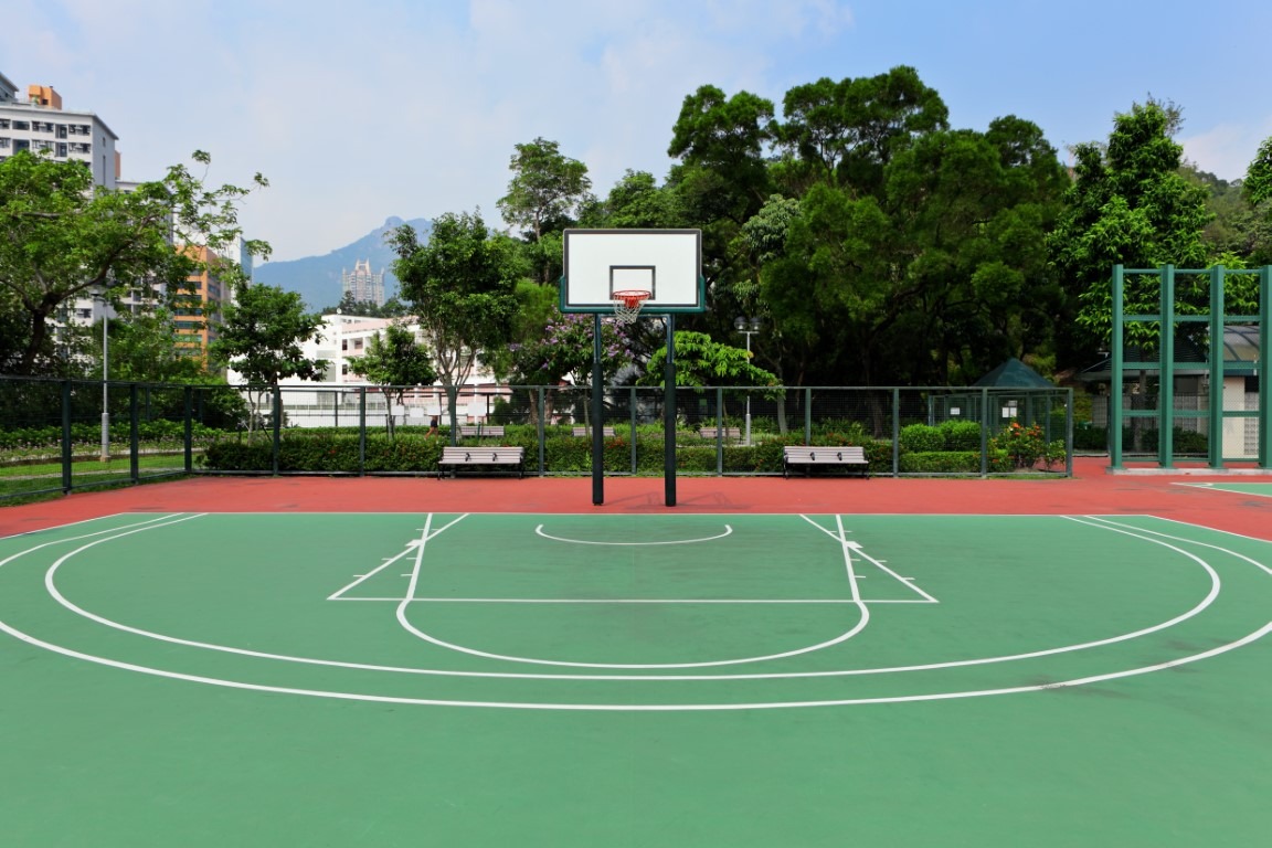 Una pista de baloncesto sin gente, con el suelo de color verde. La cámara enfoca a una canasta, con árboles tras de ella.