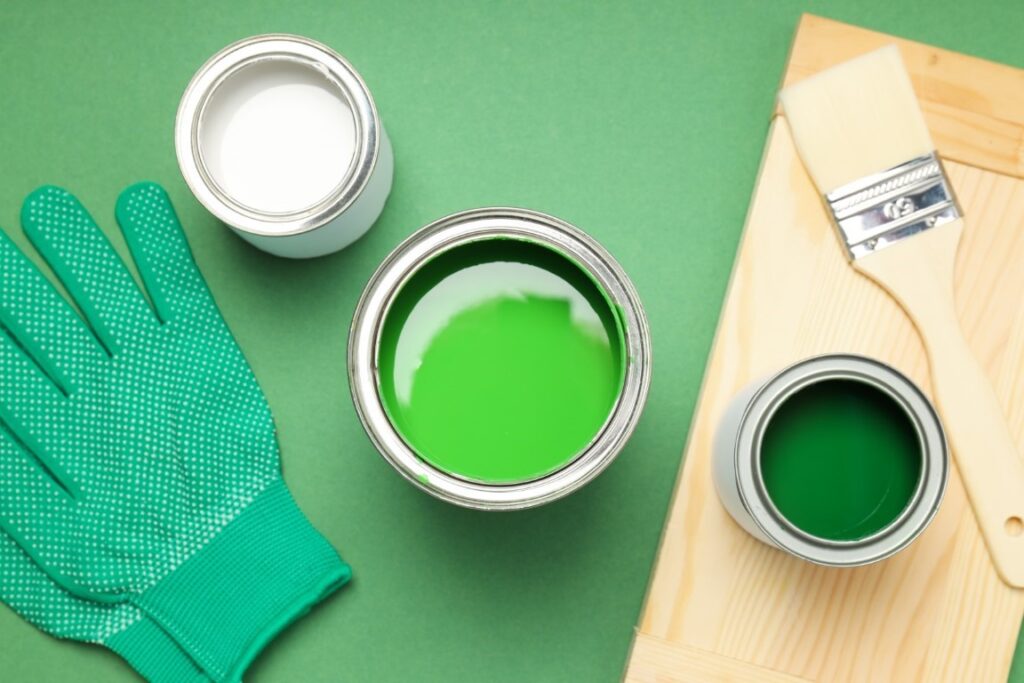 Una imagen de una superficie verde sobre la que reposan un pincel, unos guantes y tres latas de pintura