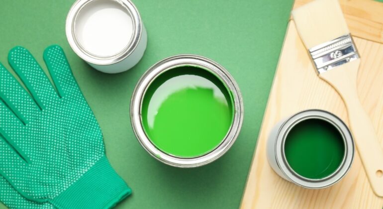 Una imagen de una superficie verde sobre la que reposan un pincel, unos guantes y tres latas de pintura