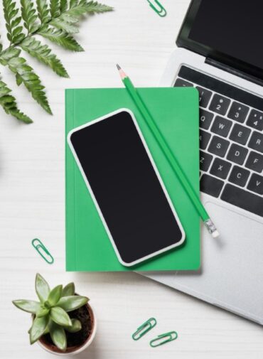 Vista cenital de una mesa en la que reposa un teléfono, un lapicero, un ordenador y un cuaderno verde