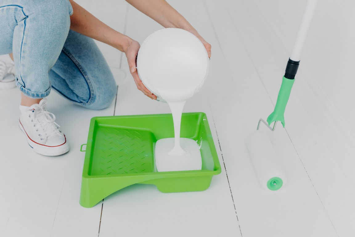 Una mujer vuelca un cubo de pintura blanca sobre un cubo verde junto a un rodillo para pintar una habitación